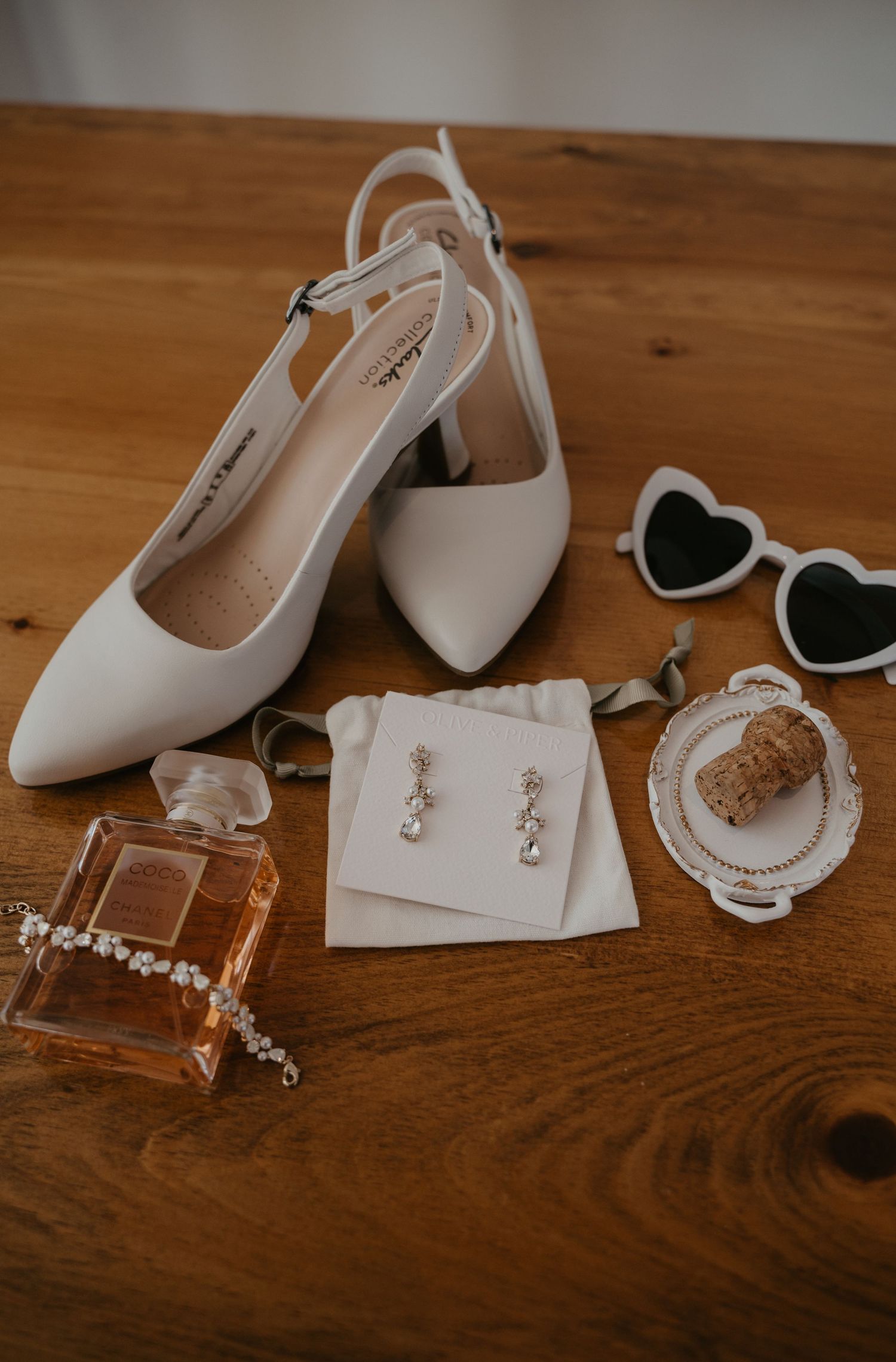 Wedding accessories including white heels, heart sunglasses, perfume, and delicate jewelry arranged on wooden surface.