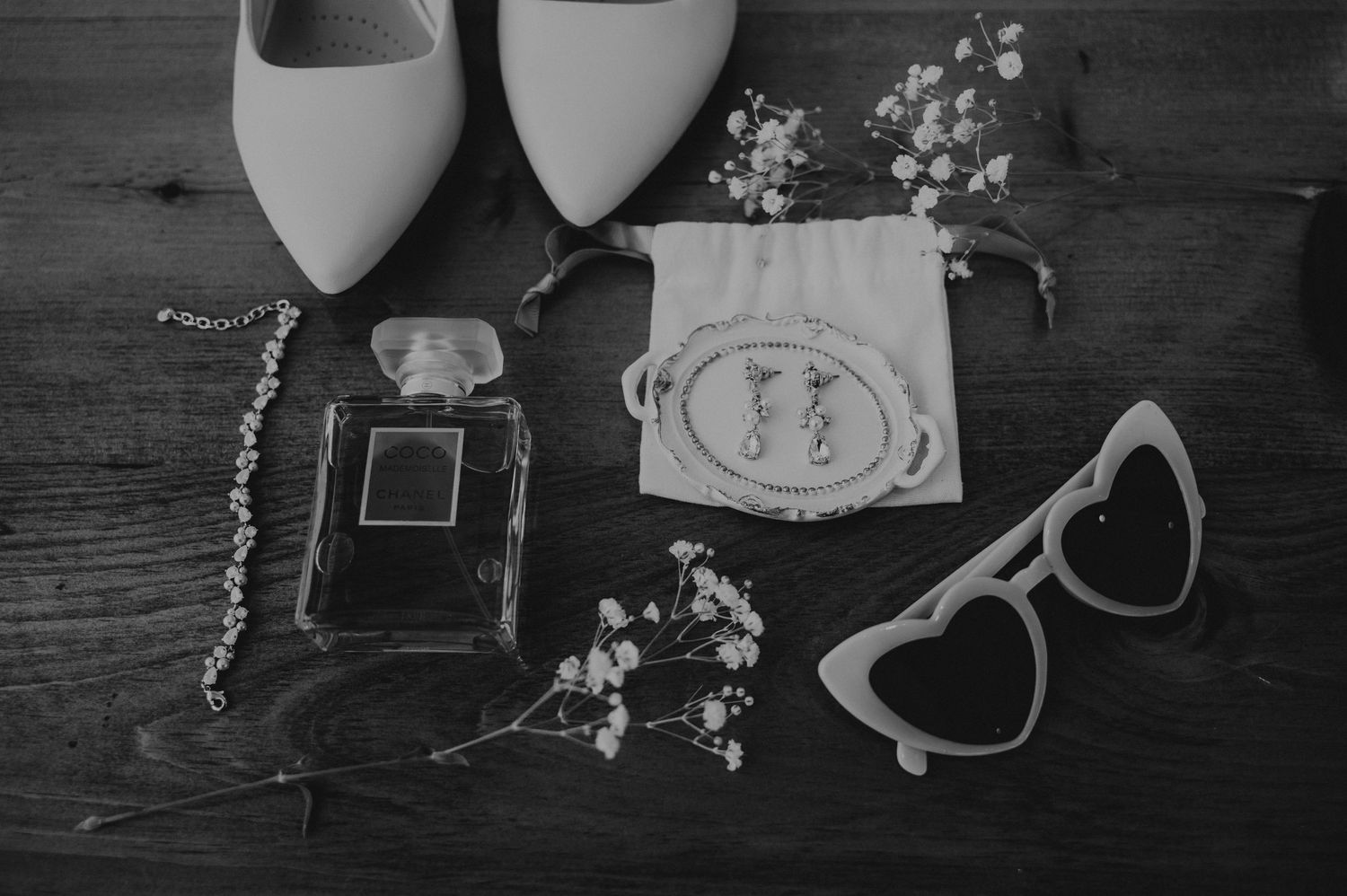 Wedding accessories including white shoes, perfume bottle, heart sunglasses and delicate flowers on dark wood surface.