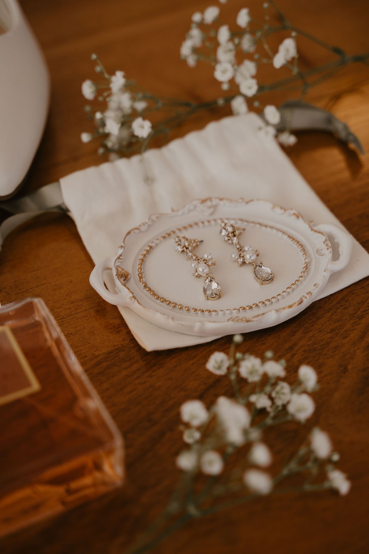 Delicate bridal jewelry and earrings displayed on white fabric with baby's breath flowers on wooden surface.