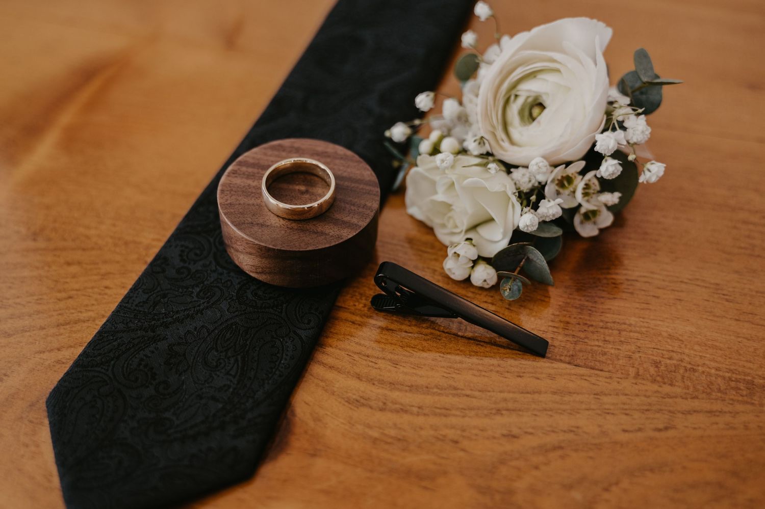 Wedding details including a black tie, wooden rings, and a delicate white floral boutonniere on wooden surface.