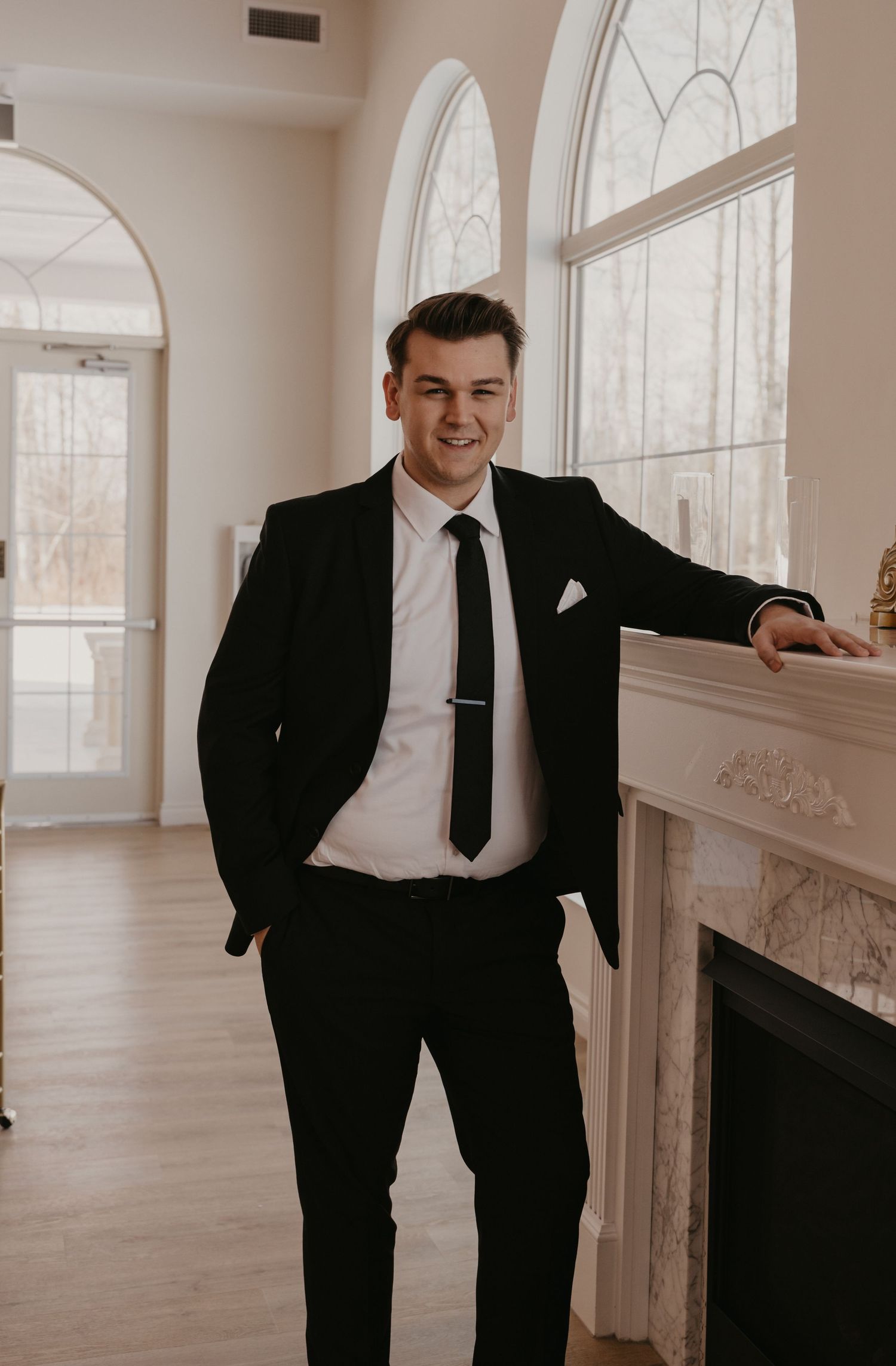 Classic black suit and tie ensemble in elegant room with arched windows and fireplace.
