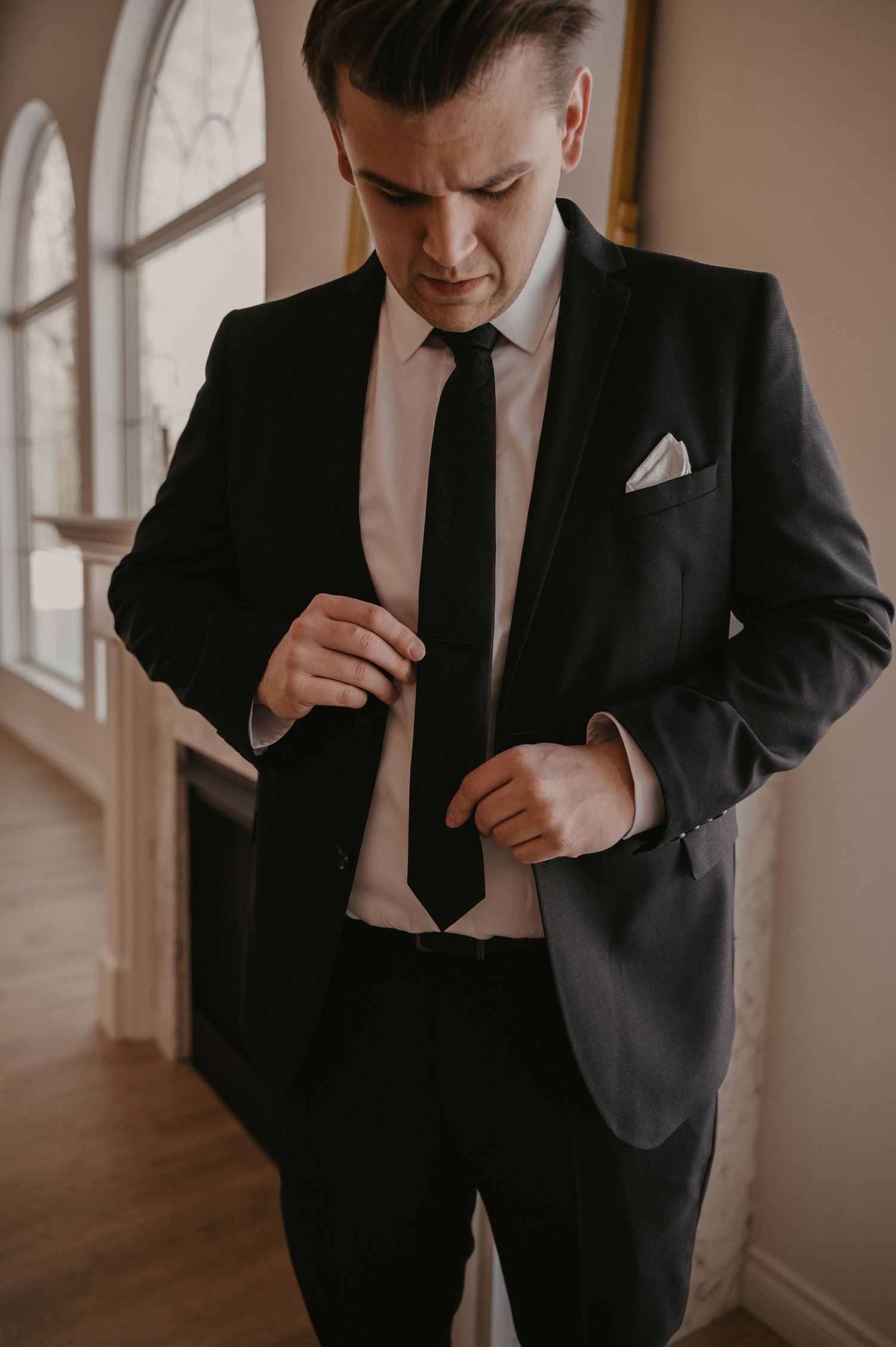 Formal black suit with tie and pocket square being adjusted in elegant setting.