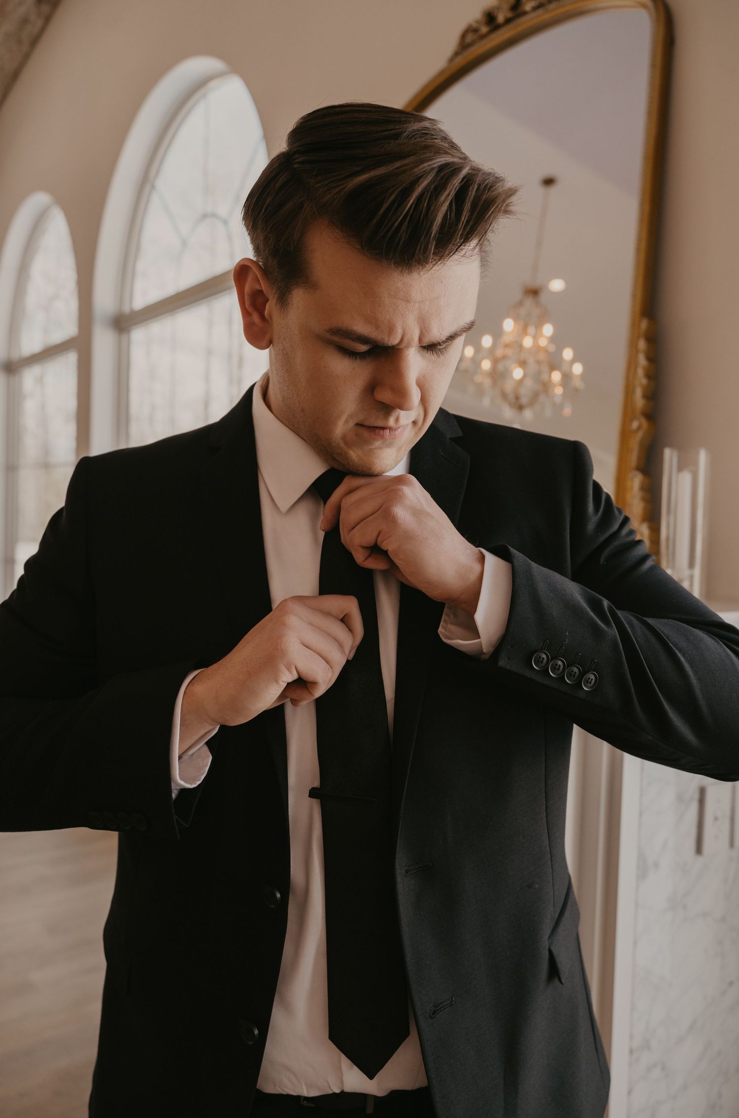 Groom adjusts his tie and suit in an elegant venue with arched windows and chandeliers.