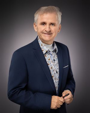 Professional studio portrait of someone wearing a navy suit jacket with floral patterned shirt against a gradient background.