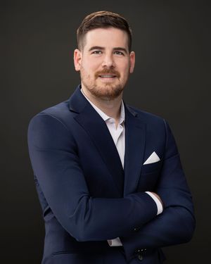 Professional studio portrait of someone in a navy suit and white dress shirt against a dark background.