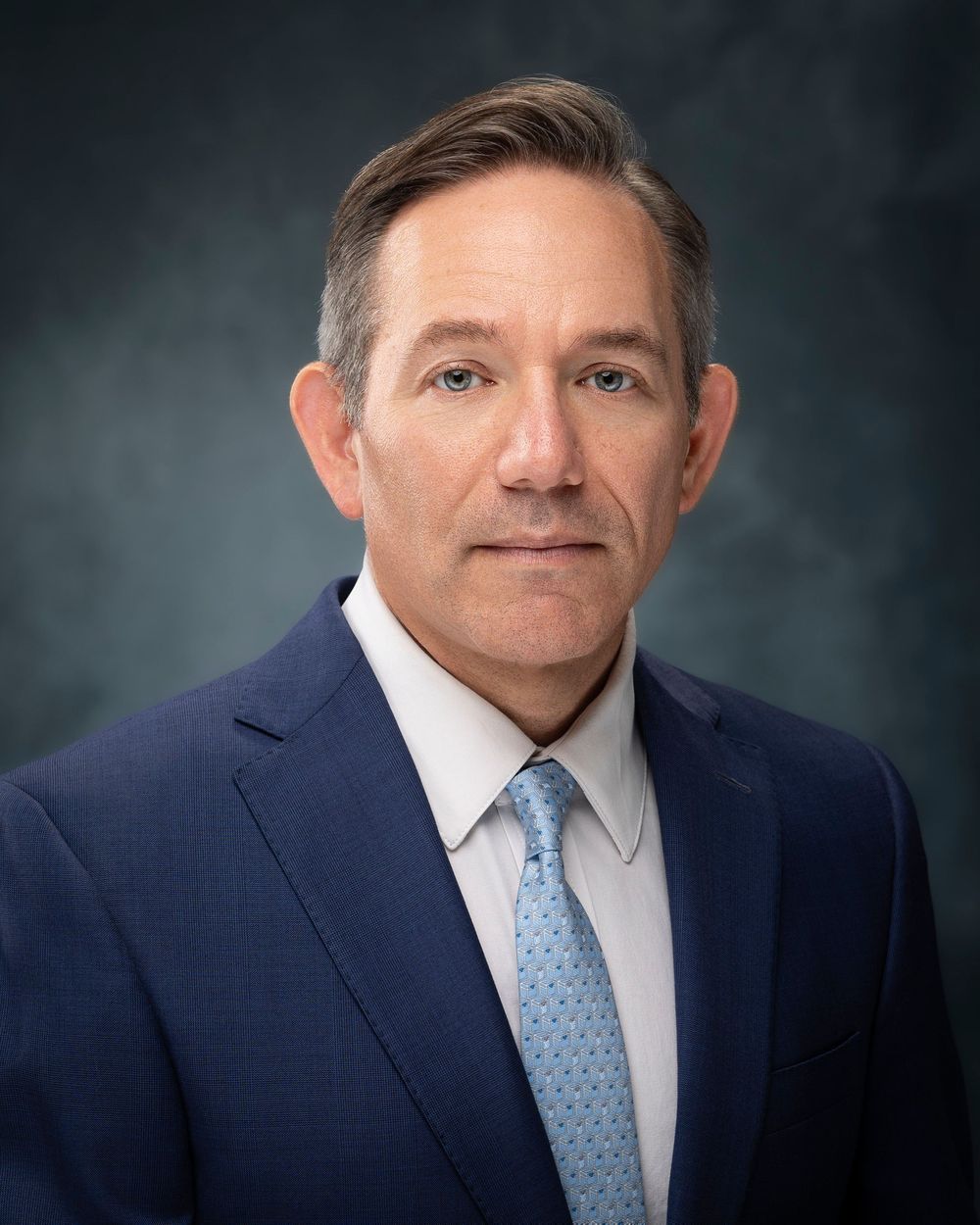 Professional headshot portrait of someone in a navy suit and light blue patterned tie against a dark background.