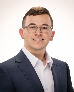 Professional headshot of someone in a dark suit jacket and white dress shirt wearing glasses against a light background.