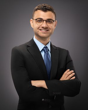 Professional business portrait of person in dark suit and blue tie against gray background.
