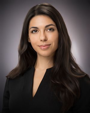 Professional headshot portrait against gray studio backdrop wearing black blazer with long dark hair.