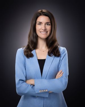 Professional portrait of someone in a light blue blazer and black top standing with arms crossed against dark background.