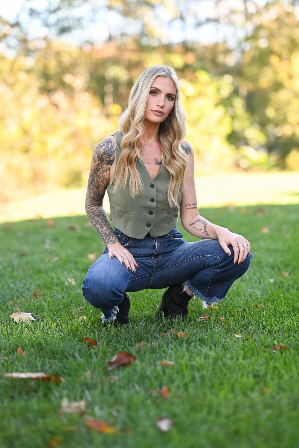 Person in olive vest and jeans poses on autumn grass with fallen leaves.