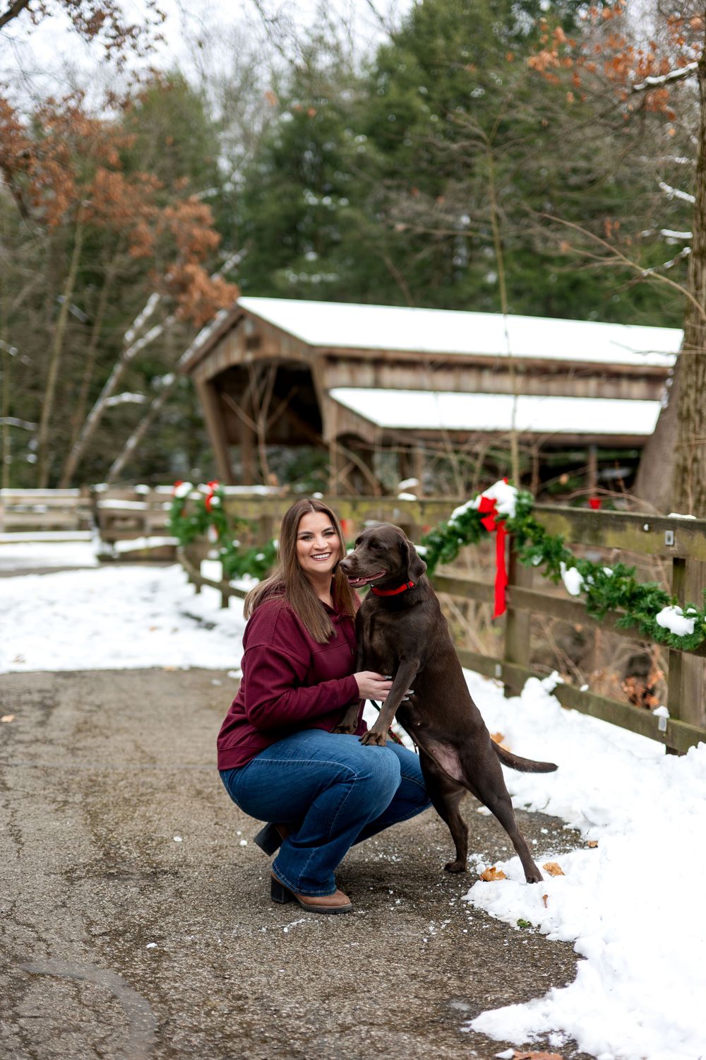 Smiling woman crouching with chocolate Lab near Christmas-decorated covered bridge in snow.