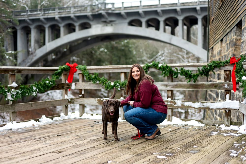 Woman crouches with chocolate Lab on snowy wooden bridge decorated with Christmas garland and bows.