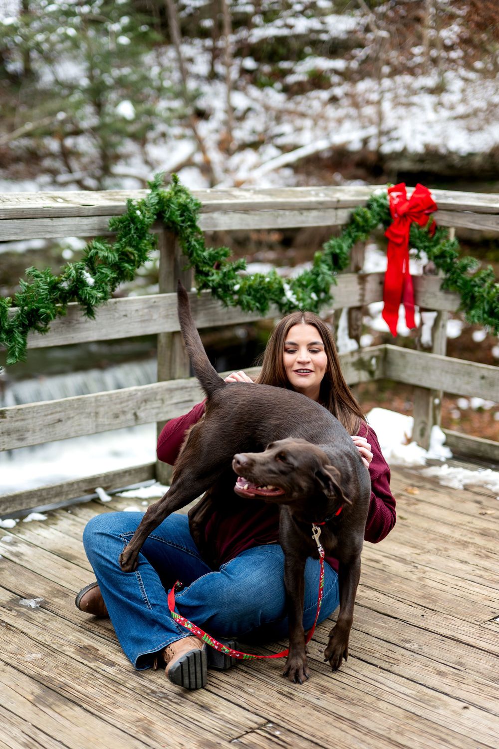 Woman in burgundy sweater hugs playful chocolate Lab on snowy Christmas decorated bridge.