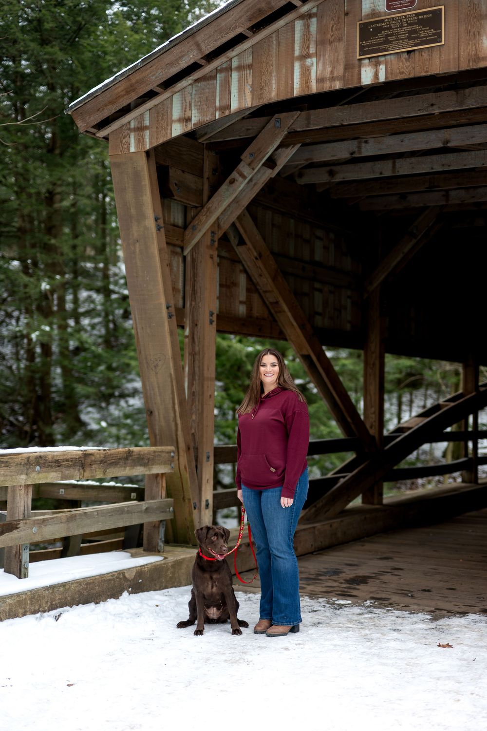 Woman standing with chocolate Labrador beside rustic wooden covered bridge in winter snow.