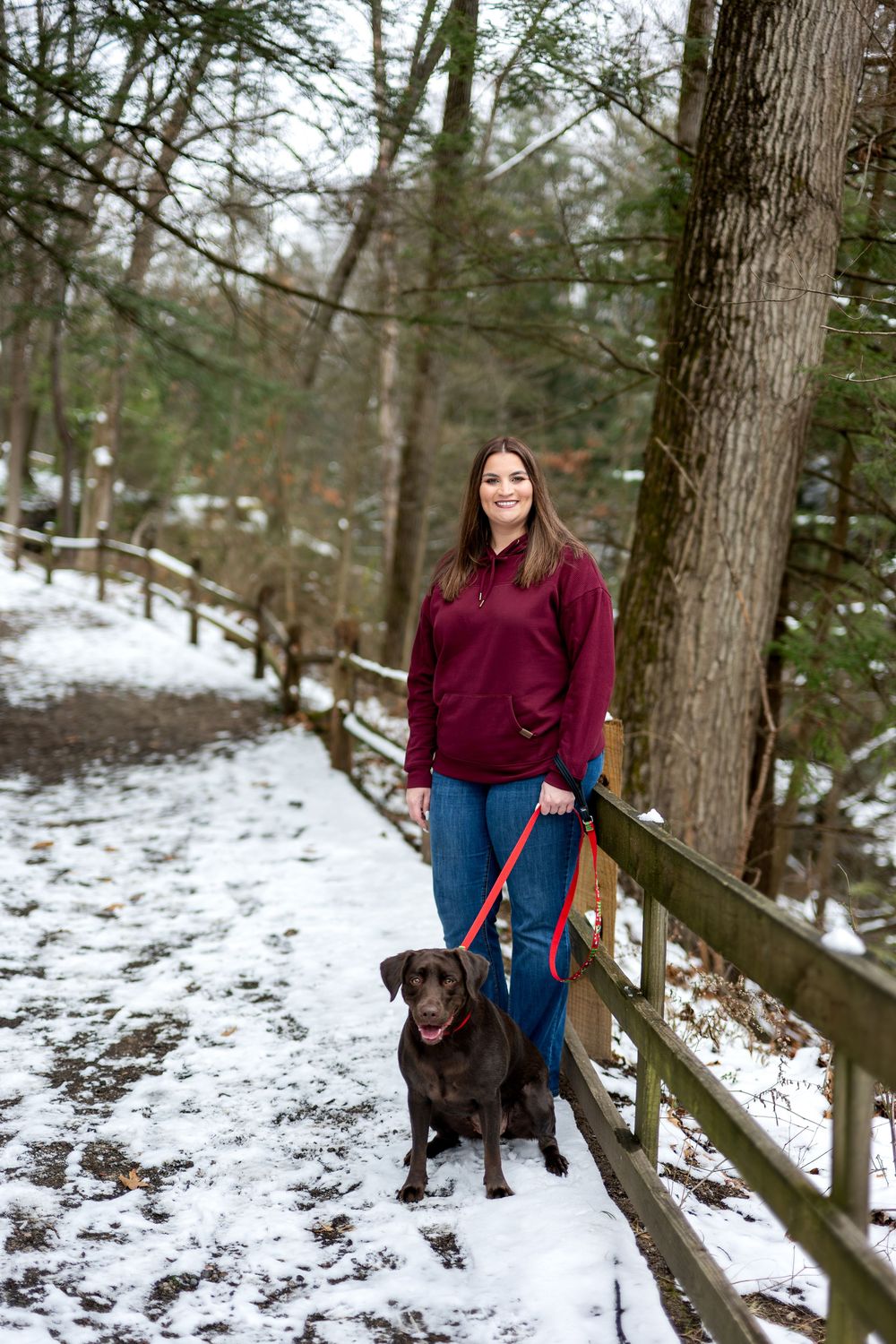 Woman in burgundy top walking chocolate Lab on red leash along snowy wooded path.