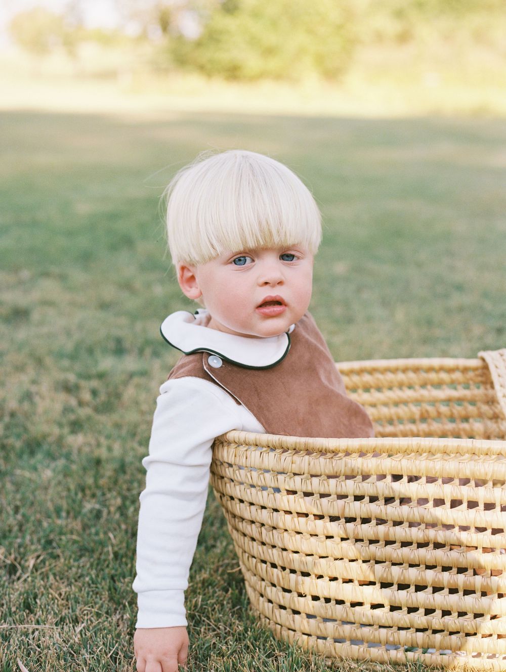 Small child in a brown and white outfit peeks out from beside a woven wicker basket on grassy lawn.