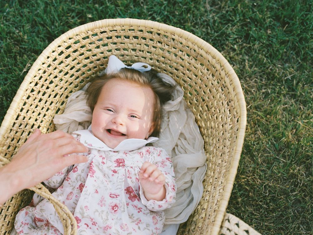 A baby in a floral dress lies on a woven basket chair in the grass, showing various happy expressions.