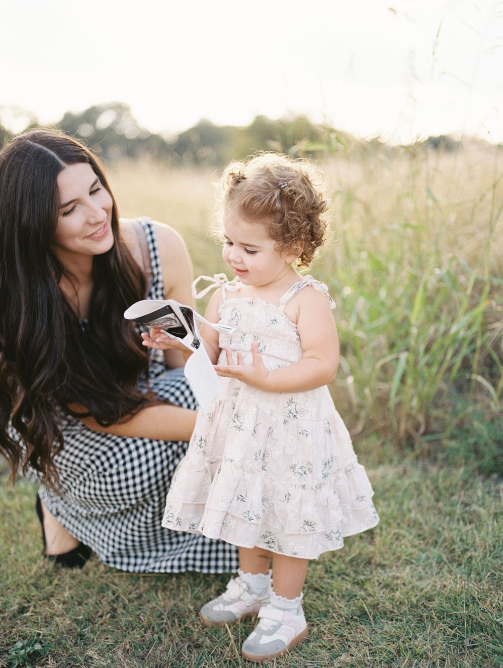 A parent and child share a tender moment together sitting in a sunlit field of tall grass and wildflowers.