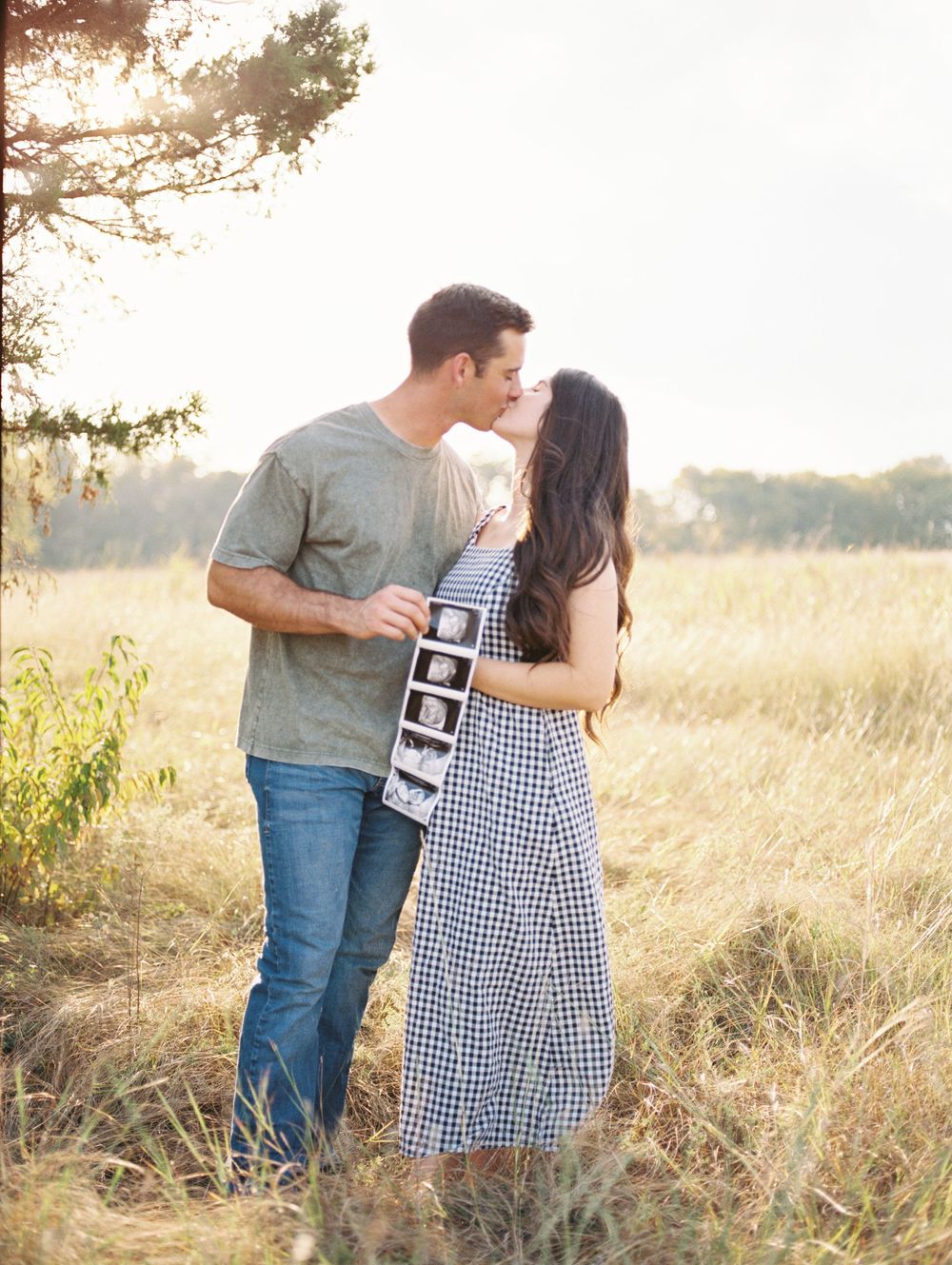 A couple embraces in a sunlit field while holding ultrasound photos during a pregnancy announcement photoshoot.