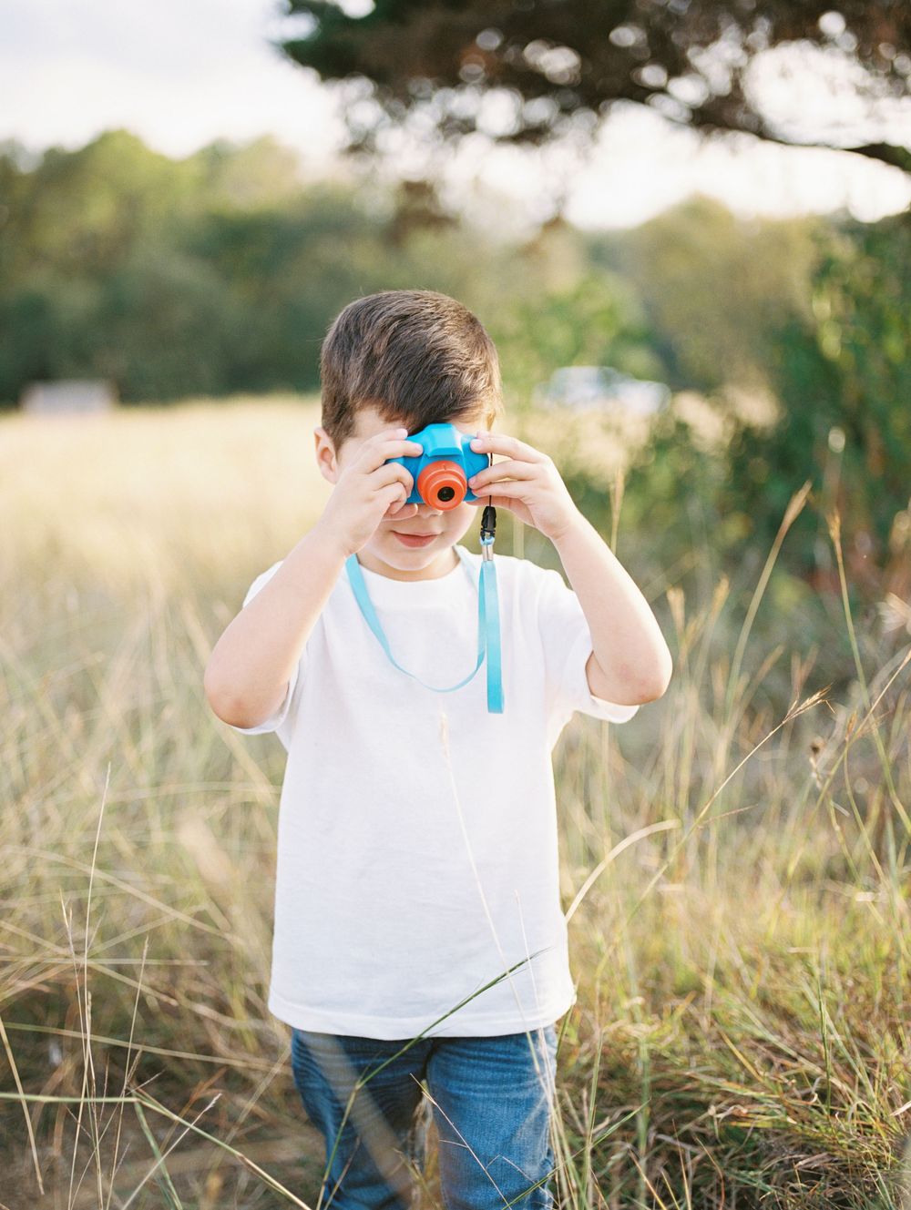 A young child plays with a blue ribbon while standing in a tall grass field at golden hour.
