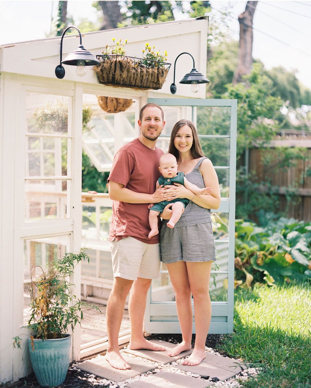 A couple stands together in a sunny garden setting with white greenhouse structures and potted plants.
