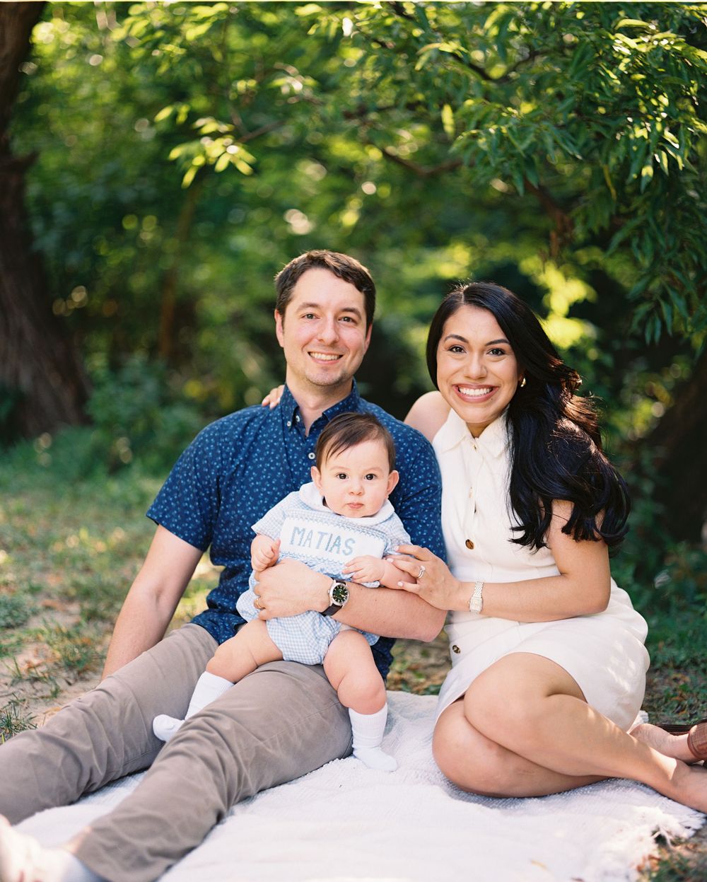 A family wearing blue and white outfits relaxes together on a white blanket in a sunlit outdoor setting.