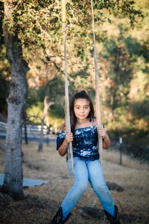 A child in blue jeans and navy shirt sits on a wooden rope swing in a sunny outdoor setting among the oak trees in her front yard home.