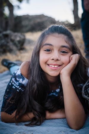 A young girl lies on the ground in a black lace dress while smiling at the camera during an outdoor photoshoot. in calaveras county