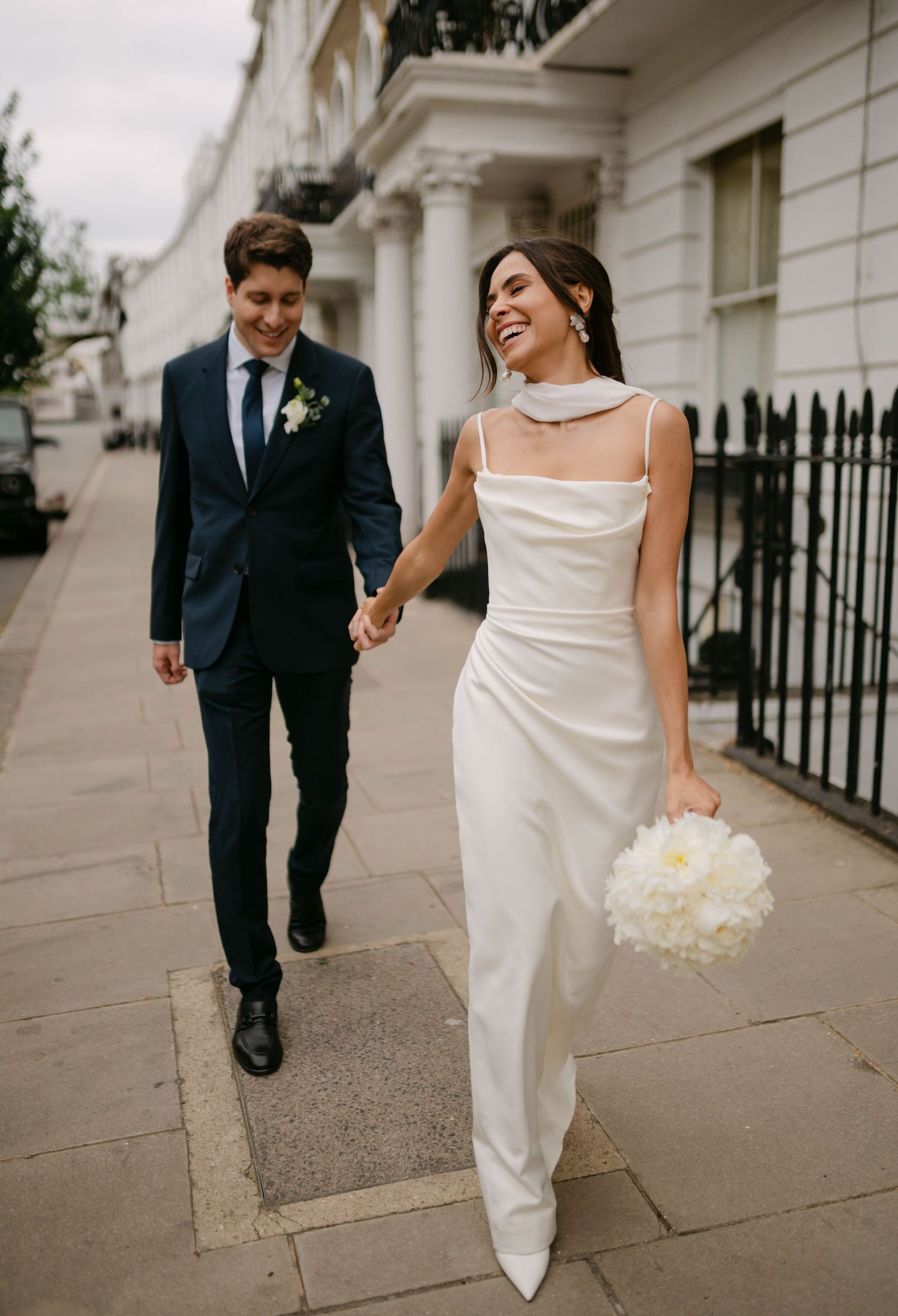 Bride in sleek white dress and groom in navy suit walking along elegant city sidewalk.