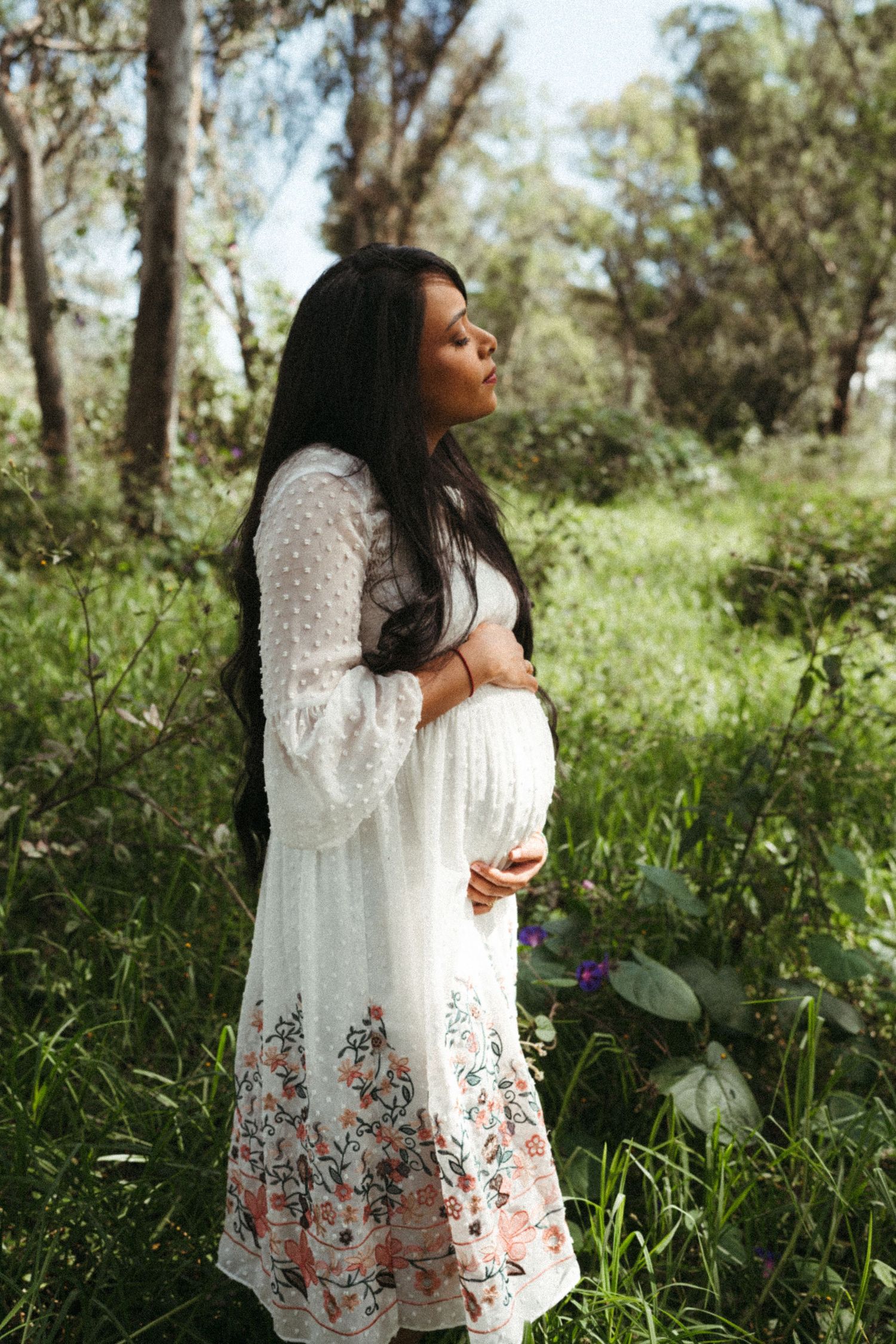 Side view of pregnant woman in white floral dress standing in sunlit forest.