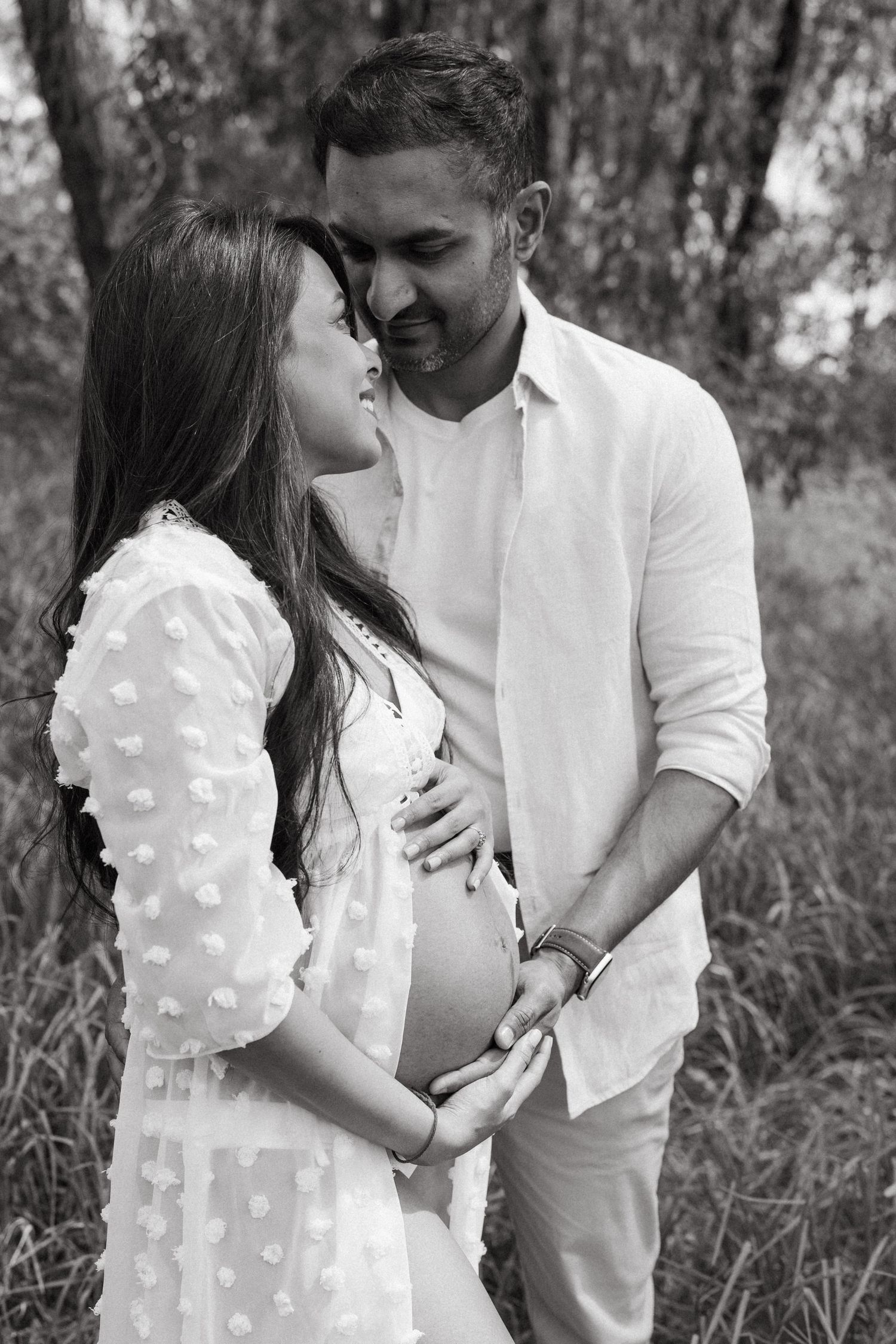 Romantic maternity photoshoot with couple in white clothing against natural backdrop.
