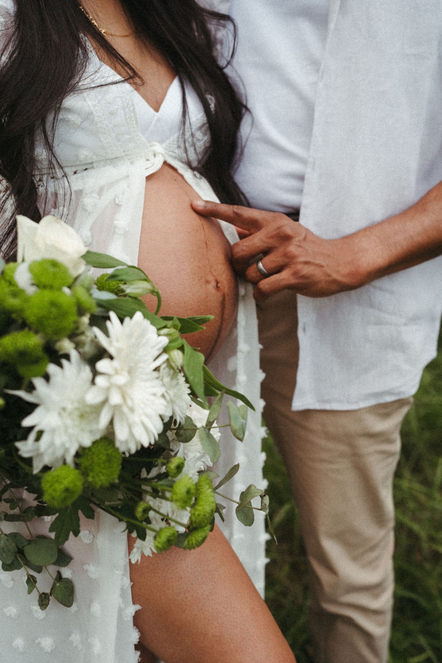 Maternity photo with white flowers and a flowing dress against a natural outdoor backdrop.