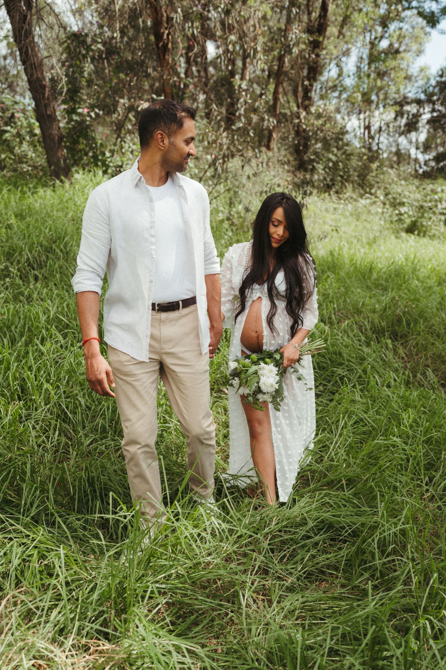 Couple walking through tall grass in white and beige outfits during a maternity photoshoot.