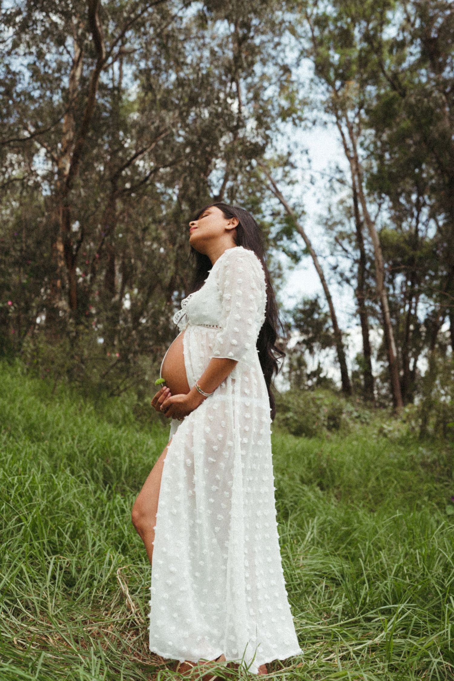 Maternity photo in a white lace dress surrounded by eucalyptus trees and natural scenery.