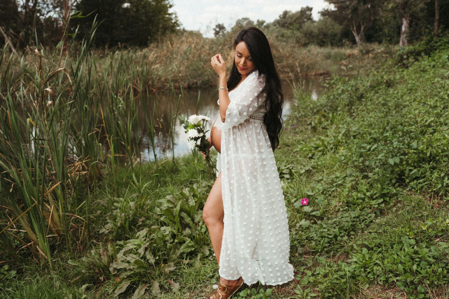 Outdoor maternity photoshoot near a pond with natural greenery and water in the background.