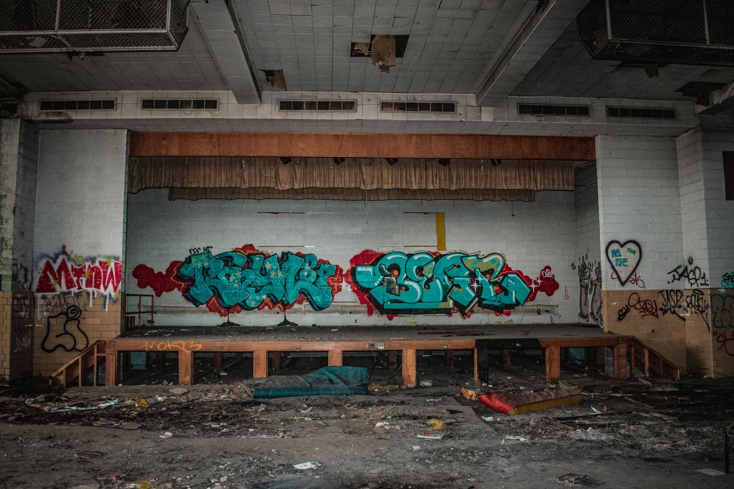 Abandoned theater with colorful graffiti murals on stage wall, debris on floor.