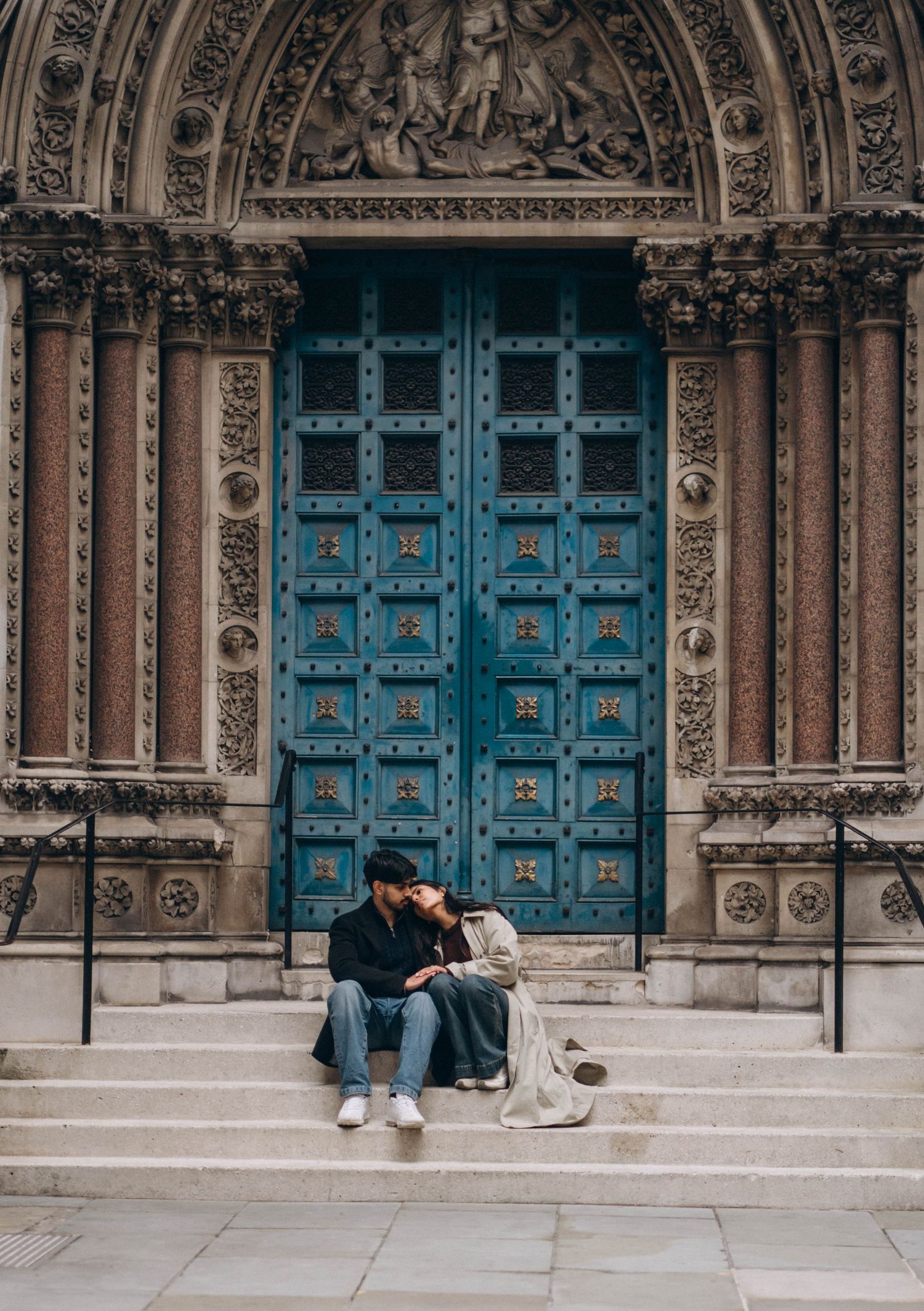 A couple sits on steps in front of ornate blue cathedral doors.