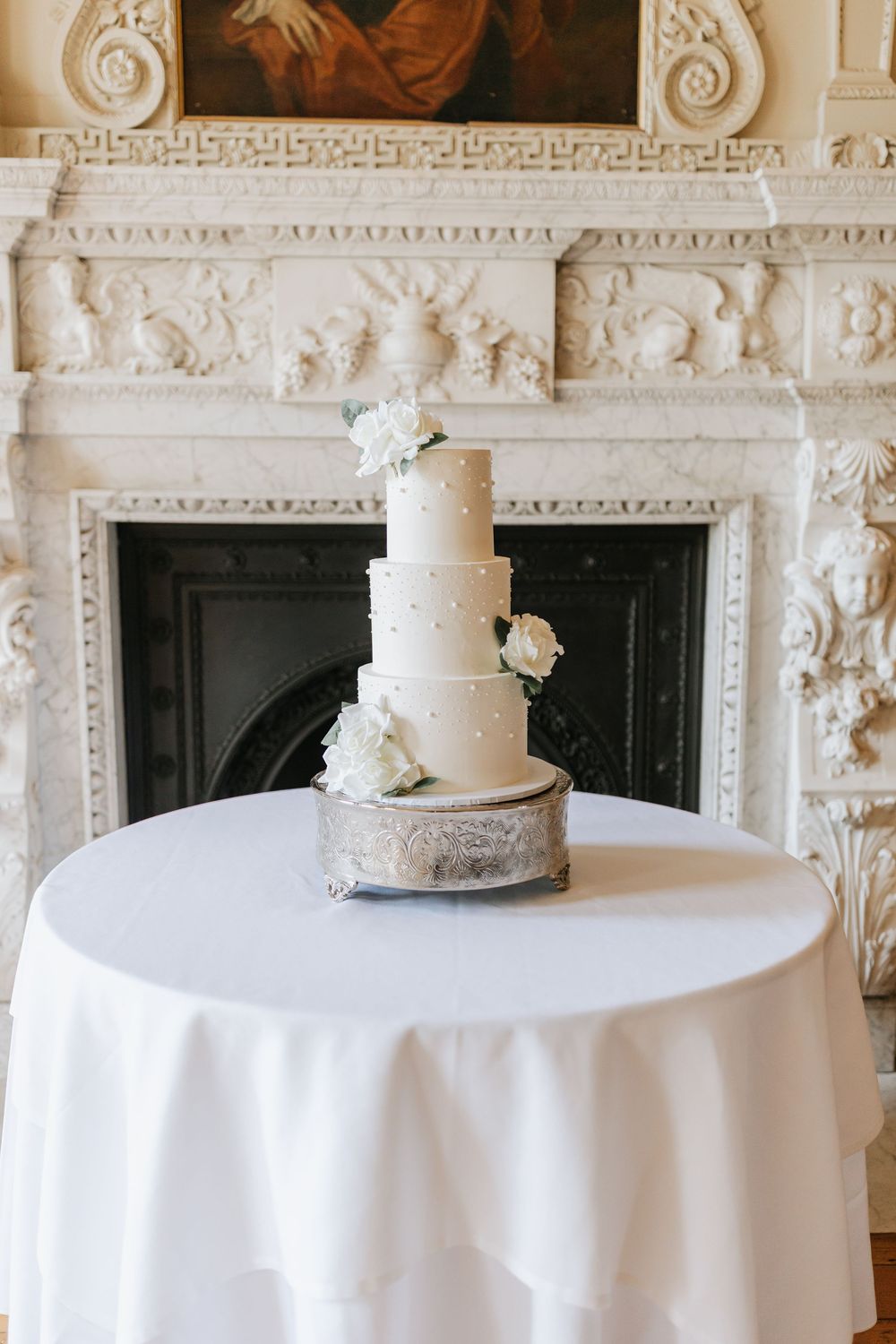 Elegant white tiered wedding cake with floral decorations displayed on round table against ornate carved fireplace mantel.