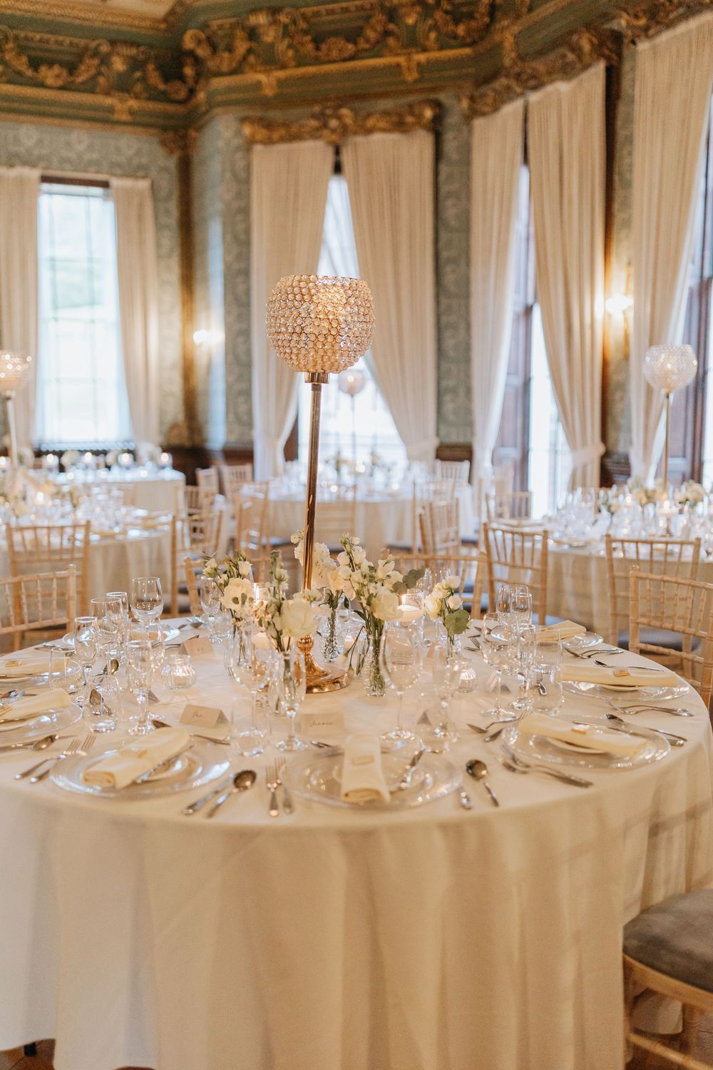 Elegant wedding reception table with crystal centerpiece, white linens and gold chiavari chairs in ornate ballroom setting.
