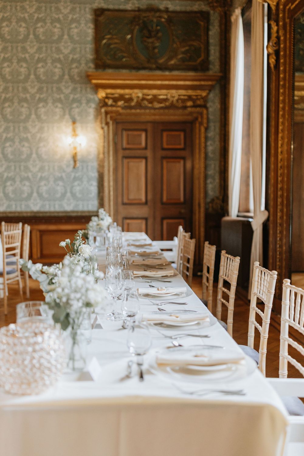 Elegant wedding reception table settings with white linens and place settings in an ornate wood-paneled dining room.