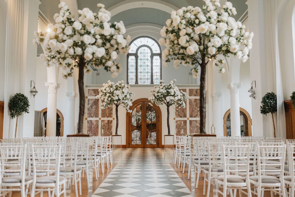 White floral arrangements line the aisle of an elegant wedding ceremony venue with arched windows and white chairs.