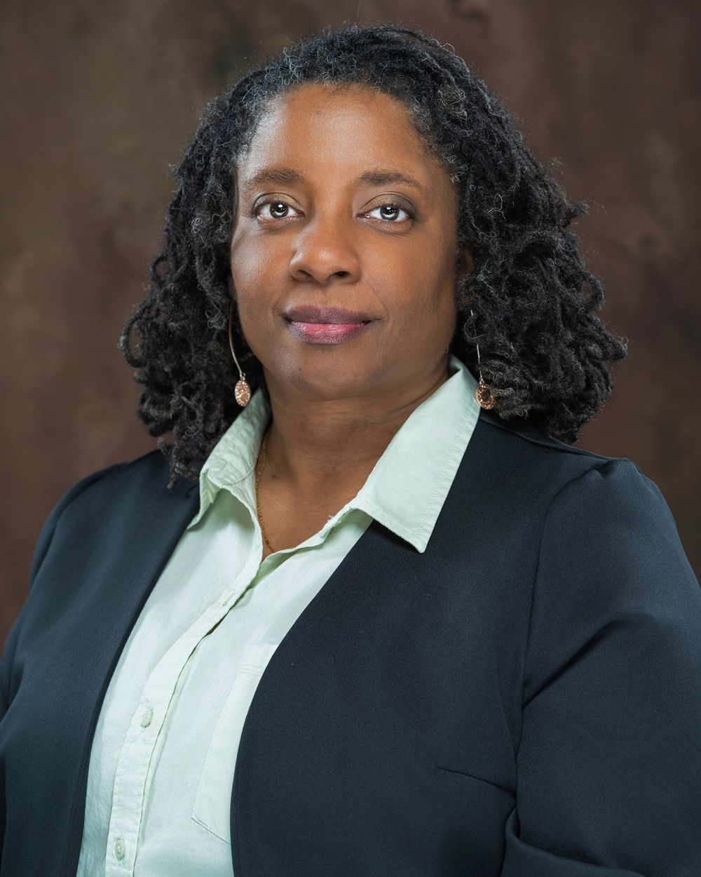 Professional headshot of a woman in a black blazer and light green shirt against a brown background.