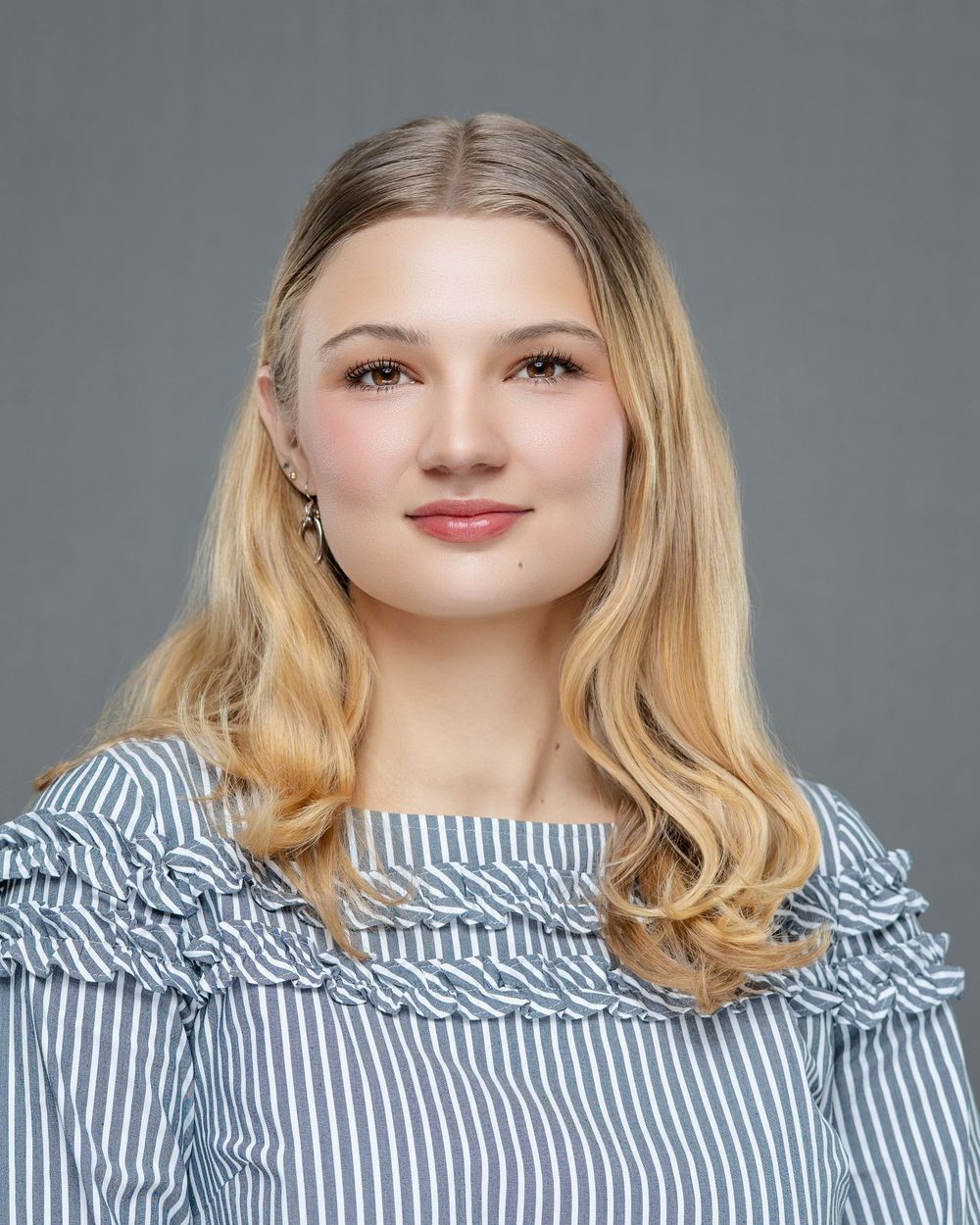 Professional studio portrait of a person with blonde wavy hair wearing a blue and white striped ruffled blouse against a gray backdrop.