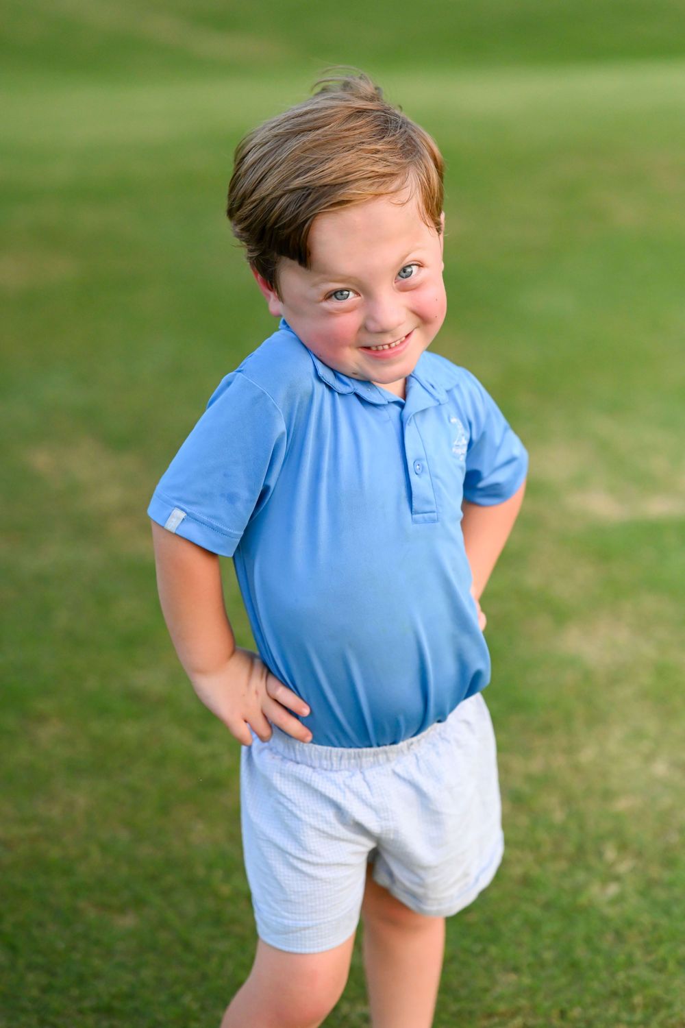 Young child in light blue polo and white shorts smiling on grass.