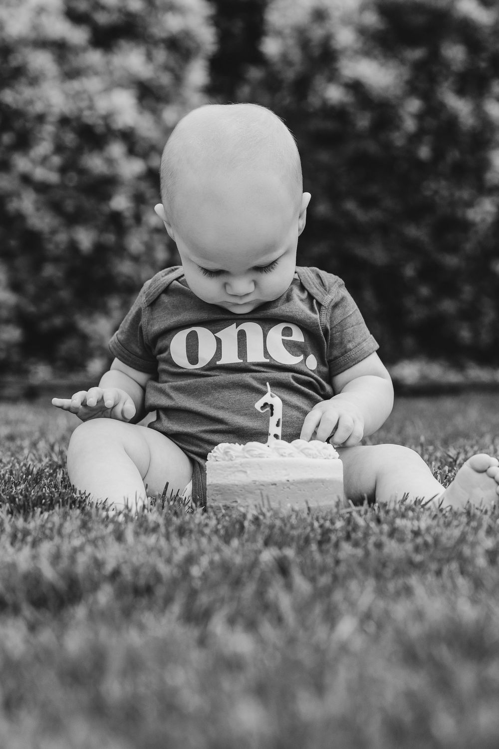 Black and white photo of a baby in a onesie sitting on grass with a small birthday cake celebrating their first birthday.