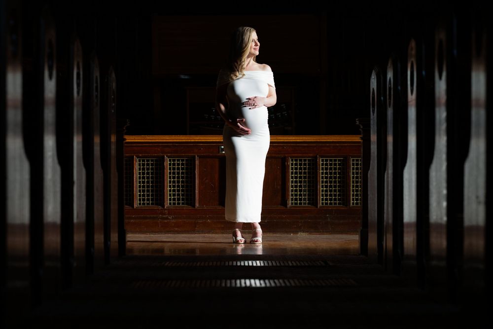 Elegant woman in white fitted dress standing in dramatic dark corridor with wooden columns.