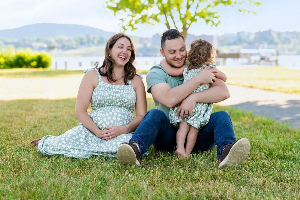 Pregnant woman in floral dress sits on grass with man hugging toddler, family maternity photo outdoors by lake.