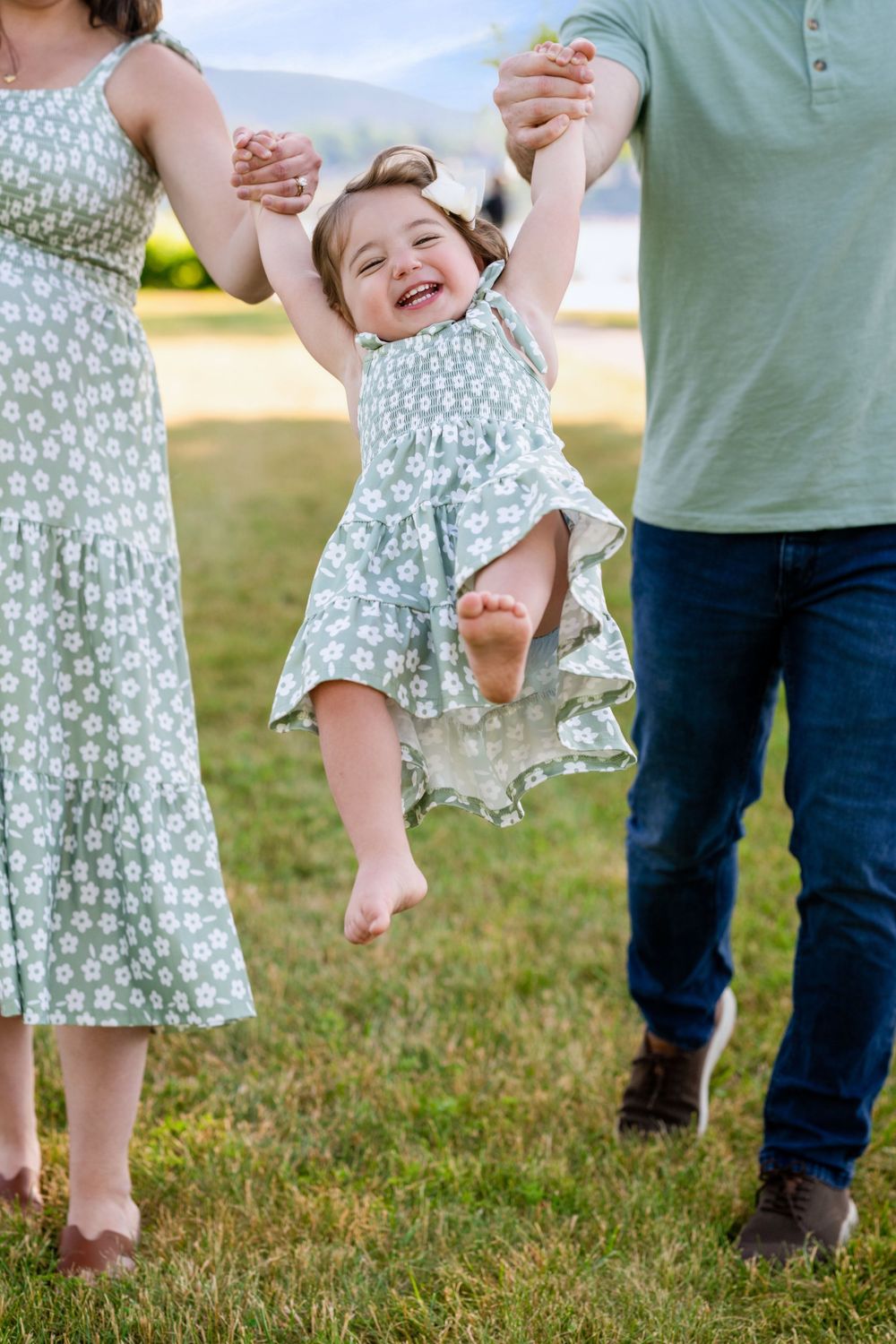 Parents joyfully swing their laughing toddler girl in a floral dress outdoors on green grass.