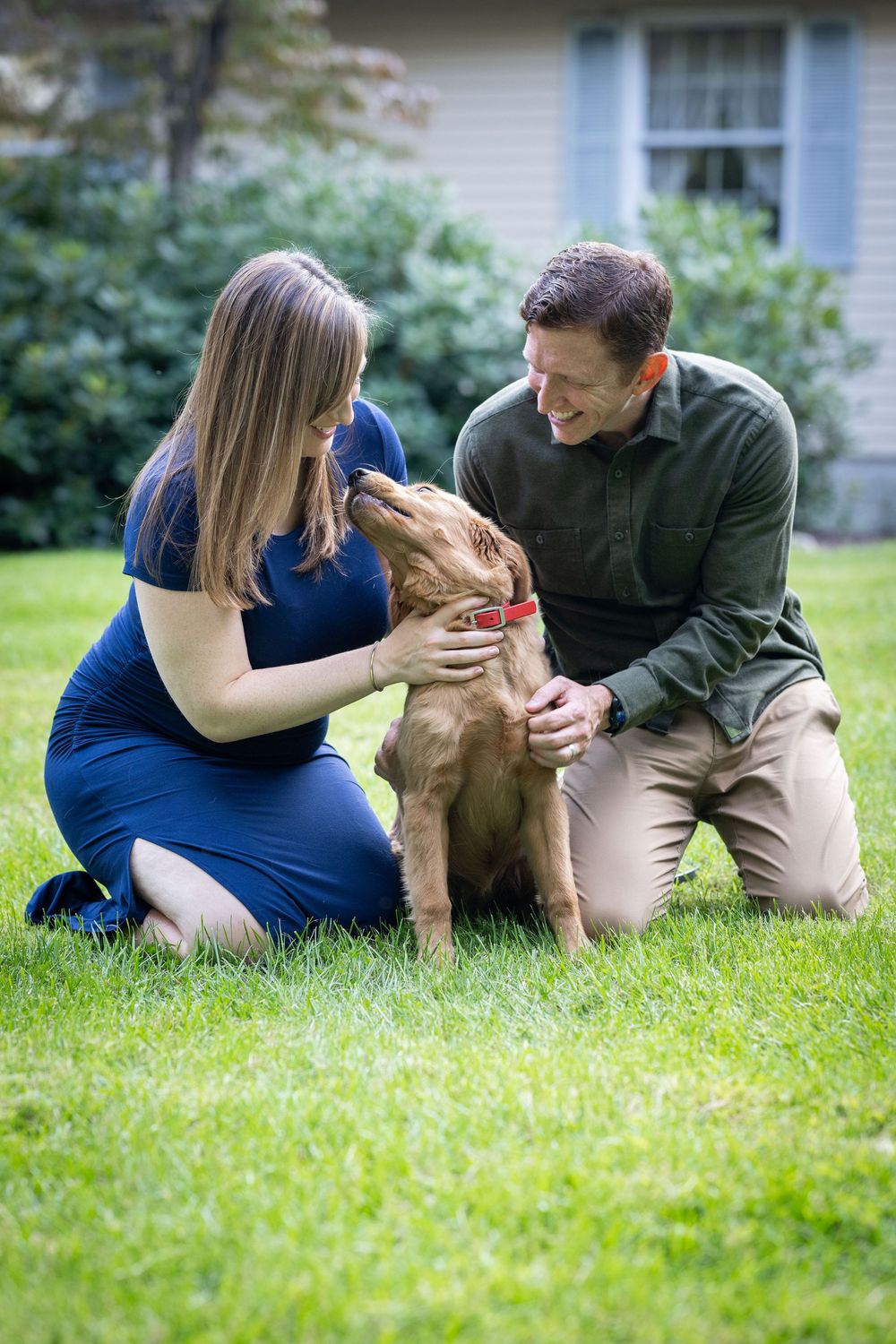 A couple kneeling on grass petting their brown dog in front of their home.